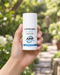 A hand holding a white bottle of APP All Purpose Peptide cream against a blurred green garden background.
